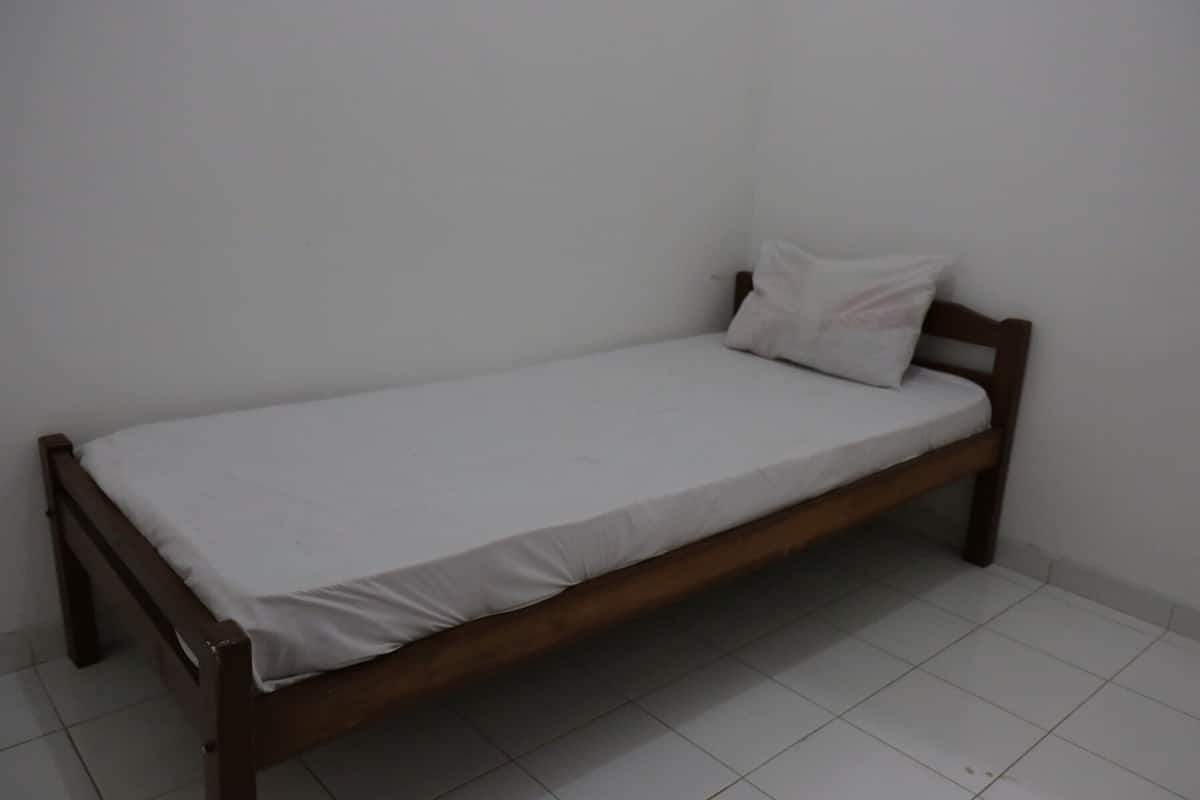 Un llit individual amb estructura de fusta i roba de llit blanca en una habitació amb parets blanques i paviment de rajoles.