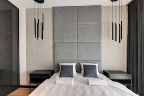 A modern bedroom with a large bed, two bedside tables, a grey padded headboard, and a hanging light fixture.