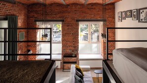 Une pièce avec des murs en briques apparentes, un lit en mezzanine, un canapé et une petite table. Il y a des fenêtres avec des rideaux et une vue sur la rue.