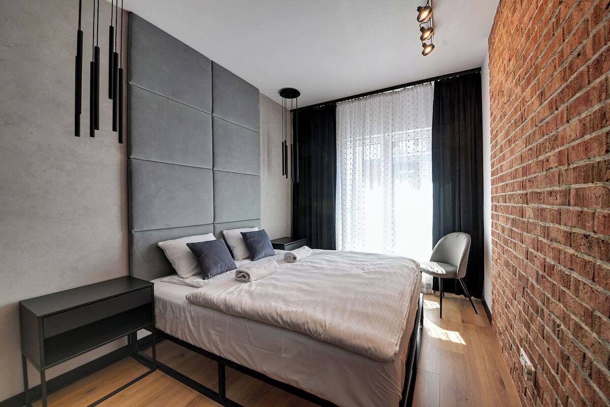 A modern bedroom with a large bed, a bedside table, a chair, and a brick wall.