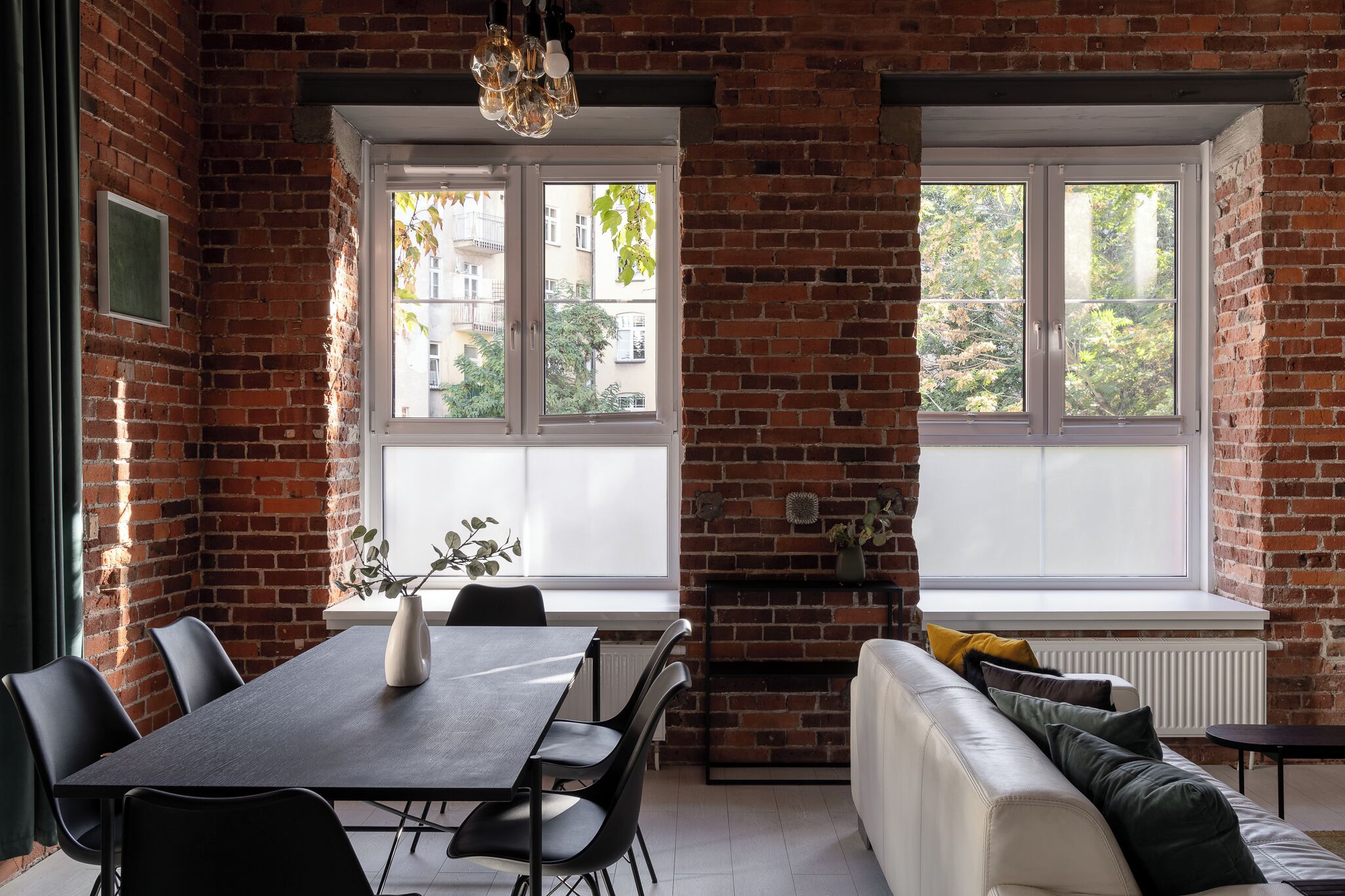 Un intérieur moderne avec un mur en briques, une table à manger avec des chaises, un canapé et deux fenêtres équipées de stores.