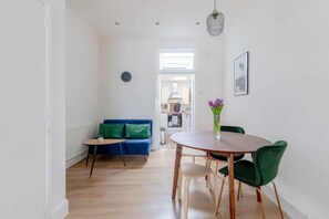 A modern living room with a blue sofa, a round wooden dining table, and green chairs. - Central & Charmingly Curated 1BD Wifi Fulham (London)