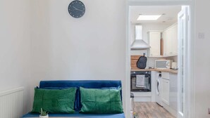 Un espacio habitable compacto con un sofá azul, una pequeña mesa redonda, un reloj en la pared y una zona de cocina con armarios y electrodomésticos blancos.