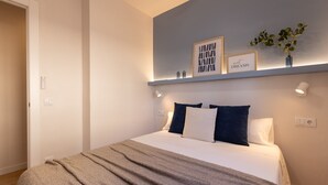Uma cama bem arrumada com um cobertor cinzento, um abajur e uma prateleira fixada na parede com quadros e uma planta.
