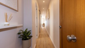 A hallway with wooden flooring, white walls, and a wooden door with a silver handle. There are framed pictures on the wall and a plant in a white pot.
