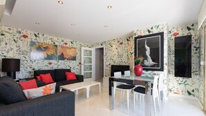 En moderne stue med sofa, sofabord og spiseområde. Der er blomstret tapet og kunstværker på væggene.