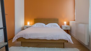 Un dormitorio con cama de madera, ropa de cama blanca, dos mesitas de noche con lámparas y una pared naranja.