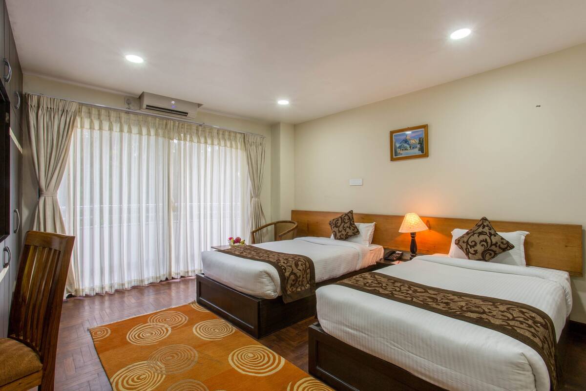 A hotel room with two beds, a wooden headboard, a nightstand with a lamp, and a large window with curtains.