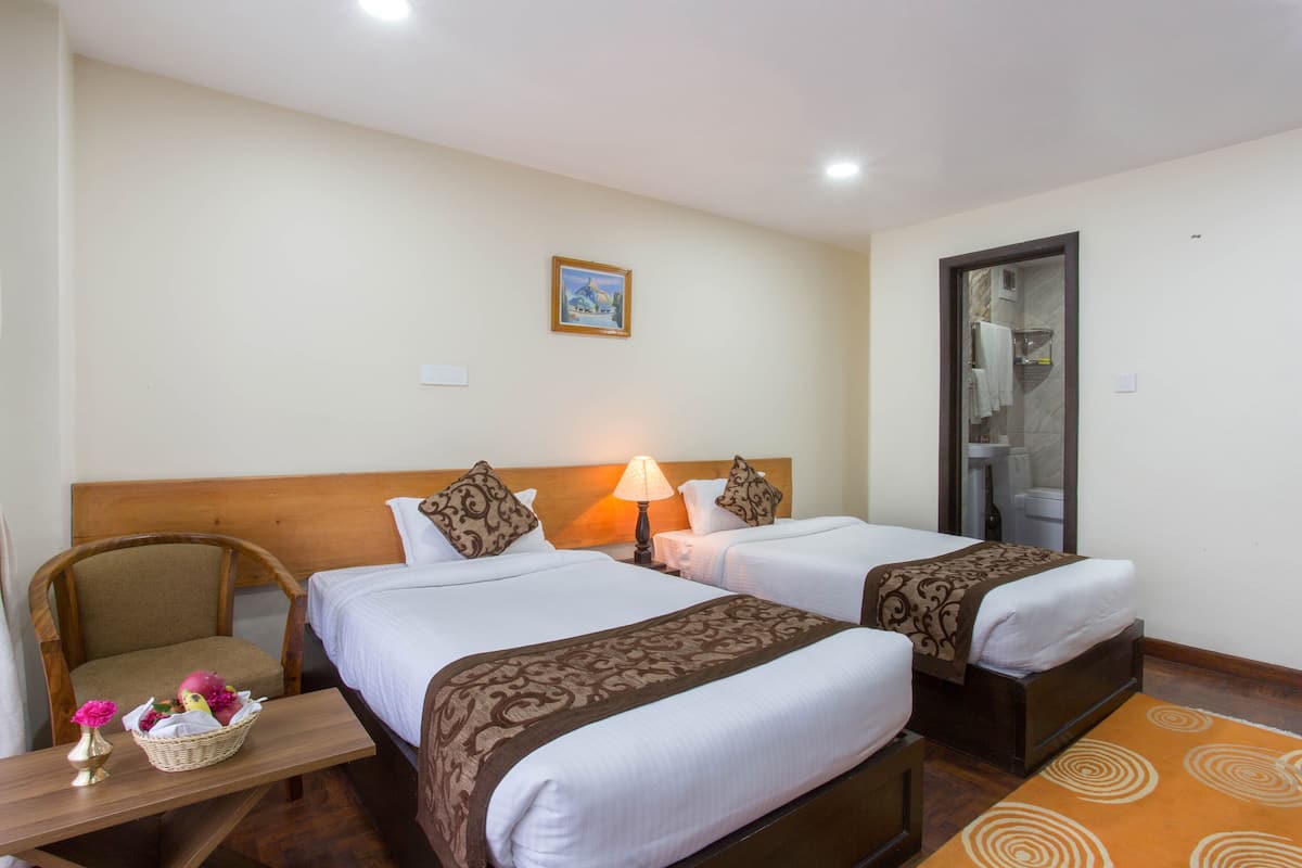 A hotel room with two beds, a wooden chair, a small table with a basket, and a bathroom visible through an open door.