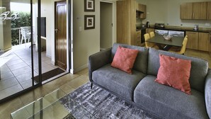 A modern living room with a grey sofa, a dining table, and a kitchen in the background. - Ellefsen Golf Suite 137, Langebaan, 4 - Sleeper (Langebaan)