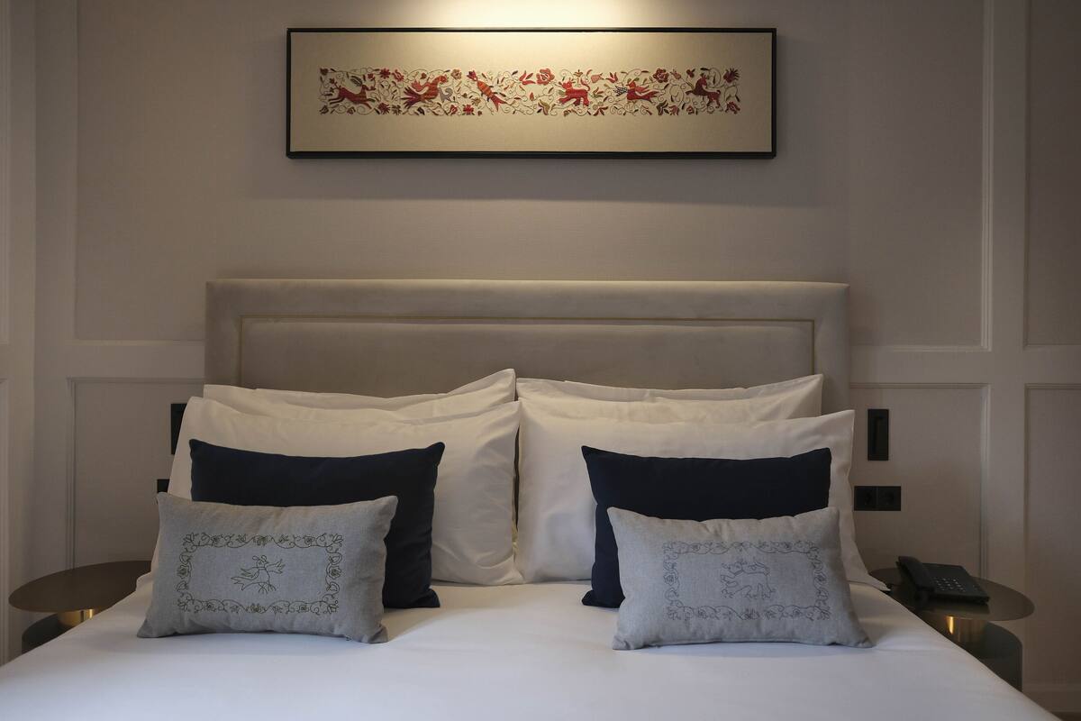 Een net opgemaakt bed met witte en donkere kussens, een lijst met een kunstwerk erboven en een nachtkastje met een telefoon.