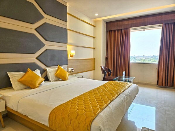 A hotel room with a large bed, a nightstand, a glass table, a chair, and a view of the city through the window.