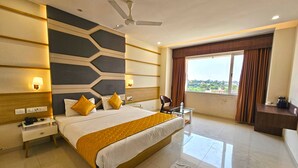Una habitación de hotel moderna con una cama grande, un ventilador de techo, una mesita de noche con lámpara, una mesa de vidrio y vistas al exterior a través de una ventana con cortinas.