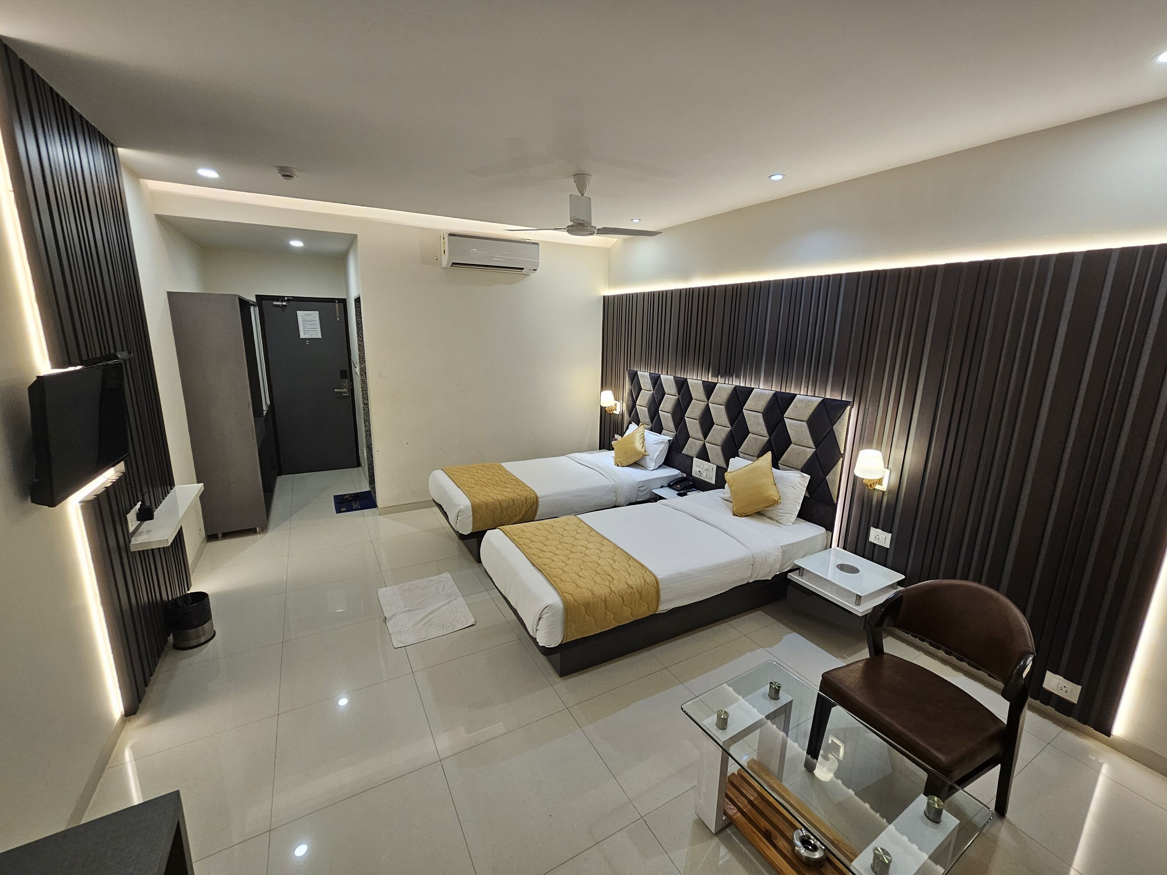 A modern hotel room with two beds, a glass coffee table, a brown chair, a TV, and a ceiling fan.