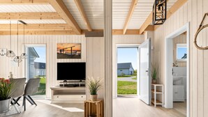 A modern living room with a flat-screen TV, wooden beams, a dining area with chairs, and a view of a house and garden. - Skippers Home - Terrace, Fireplace, Infrared Sauna, Free Pets Allowed (Olpenitz)