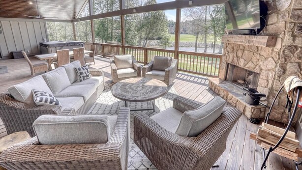 A covered outdoor patio with wicker furniture, a stone fireplace, and a television mounted on the wall. - Spacious Home in Appalachian Mountains - Hot Tub, Firepit, on the Tocccoa River! (Suches)