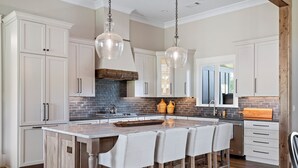 A kitchen with white cabinets, a central island with bar stools, hanging pendant lights, and a brick backsplash. - Spacious Home in Appalachian Mountains - Hot Tub, Firepit, on the Tocccoa River! (Suches)