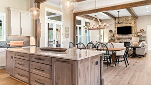 A spacious kitchen with a large island, wooden cabinets, and a stone fireplace. - Spacious Home in Appalachian Mountains - Hot Tub, Firepit, on the Tocccoa River! (Suches)