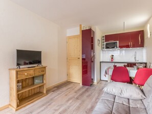 Um espaço compacto com televisão sobre suporte de madeira, uma kitchenette com armários vermelhos e uma área de refeições com cadeiras vermelhas.