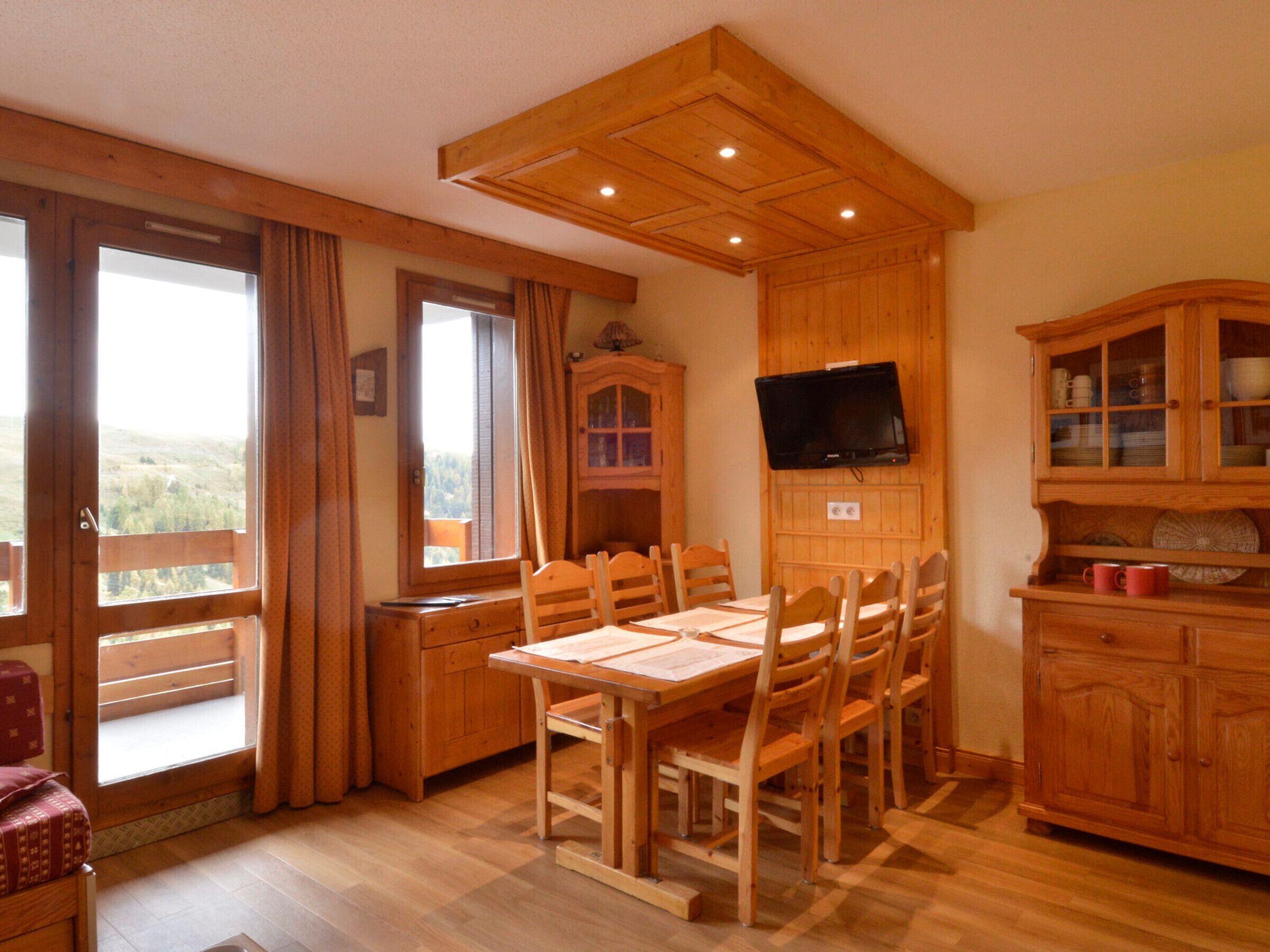 A wooden dining table with chairs, a television mounted on the wall, and wooden cabinets.
