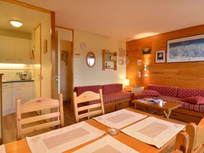 Uma sala de estar acolhedora com uma mesa de madeira, dois sofás vermelhos estampados e uma cozinha visível por uma porta aberta.