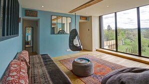 Une pièce spacieuse avec une grande fenêtre, un fauteuil suspendu, un canapé et un tapis à motifs.