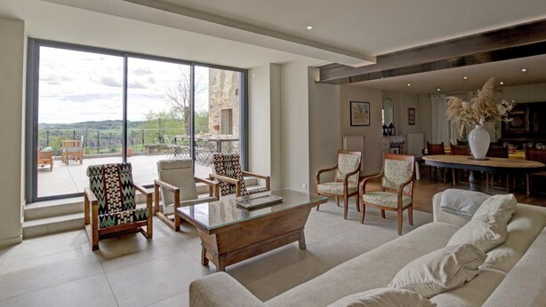 Un vaste salon doté d’une grande porte vitrée donnant sur une terrasse extérieure, aménagée avec un canapé, des fauteuils et une table basse.