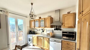 A compact kitchen with wooden cabinets, a built-in oven, a washing machine, and a dining area with a chair.