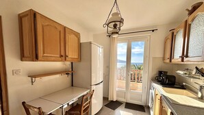 A compact kitchen with wooden cabinets, a white refrigerator, a dining table with chairs, and a view of the sea through a glass door.