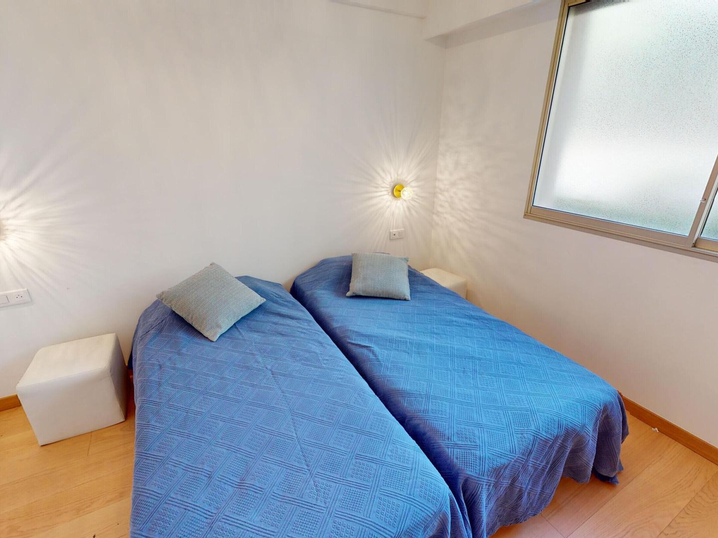 Ein Zimmer mit zwei Betten, jedes mit blauer Bettdecke und Kissen. Es gibt ein Fenster mit Milchglas, eine weiße Wand und einen kleinen, weißen Nachttisch neben jedem Bett.