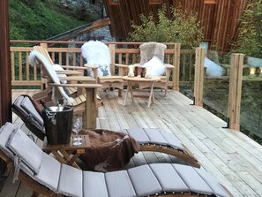 A wooden deck with lounge chairs, a table with a bottle and glasses, and a stone wall in the background.