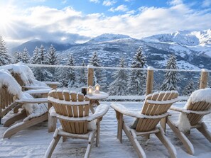 A snow-covered outdoor seating area with wooden chairs and tables, overlooking a mountainous landscape.
