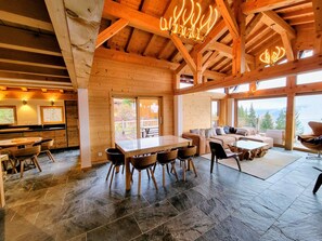 Interior espacioso de una cabaña de madera con comedor, un conjunto de sofás y ventanas amplias con vistas al exterior.