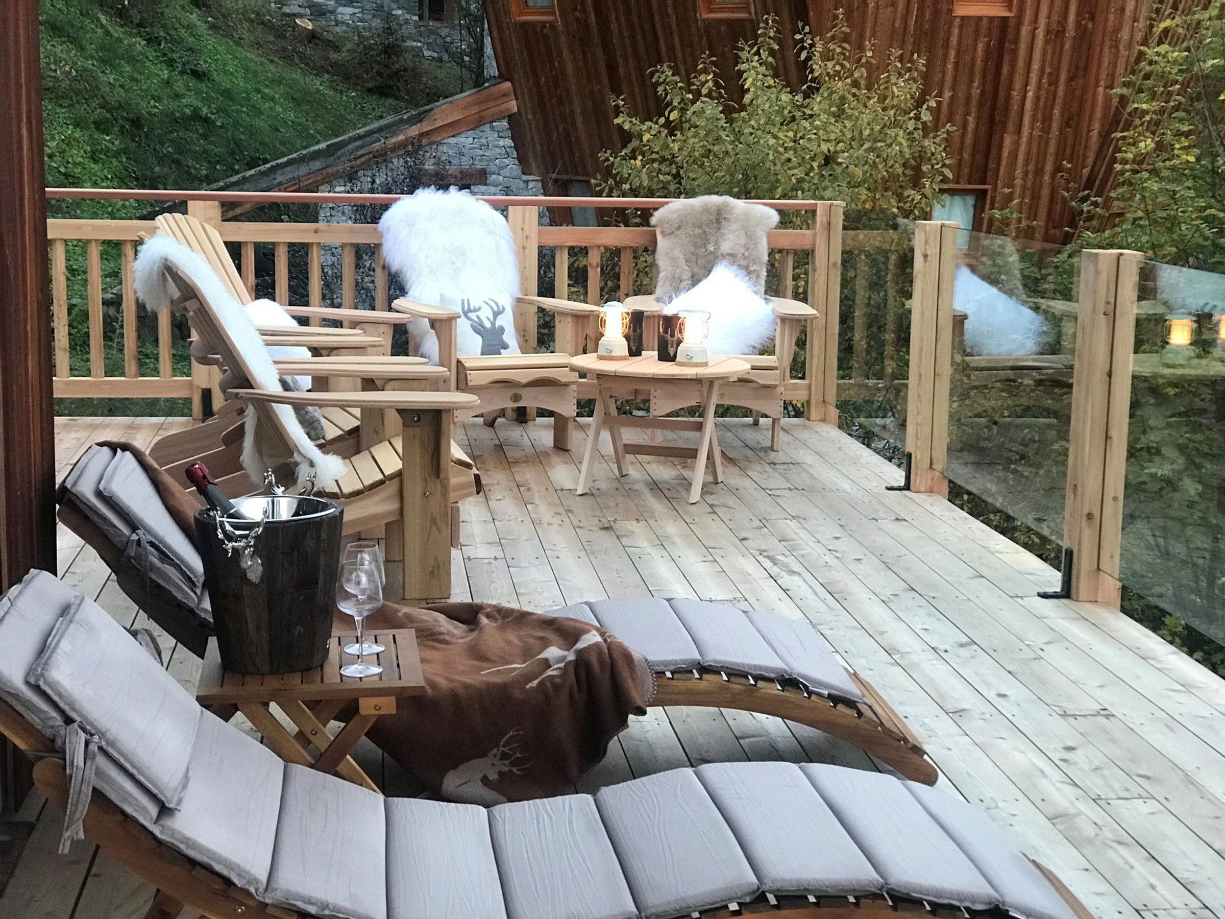 A wooden deck with lounge chairs, a table with a bottle and glasses, and a stone wall in the background.