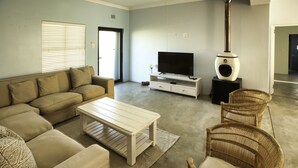 A living room with a beige sofa, a white coffee table, wicker chairs, a flat-screen TV, and a wood-burning stove. - Sandy Feet Langebaan 8-sleeper (Langebaan)