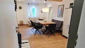 Una habitació moderna i ben il·luminada amb terra de fusta, una taula de dinar amb cadires, un sofà i una petita taula auxiliar amb un rellotge.