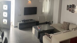 A modern living room with a sofa, coffee table, and TV. There is a door with glass panels and a window with curtains. - Mykonos Chora 2 Bd House Cycladic Escape (Mykonos)