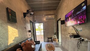 A cozy living room with a leather sofa, two large teddy bears, a small wooden table, a television mounted on the wall, and a large window providing natural light.