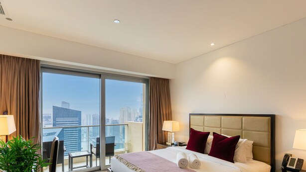 A hotel room with a large window, a bed with red pillows, a nightstand with a phone, and a view of the cityscape. - Elegant Studio Address Marina (Dubai)