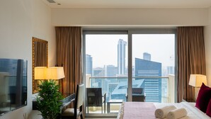 A hotel room with a large window, a bed with towels, a desk with a lamp, a chair, and a television. - Elegant Studio Address Marina (Dubai)
