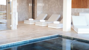 Un área de piscina interior moderna con agua azul cristalina, sillones blancos para relajarse y una zona de ducha con cerramiento de vidrio.
