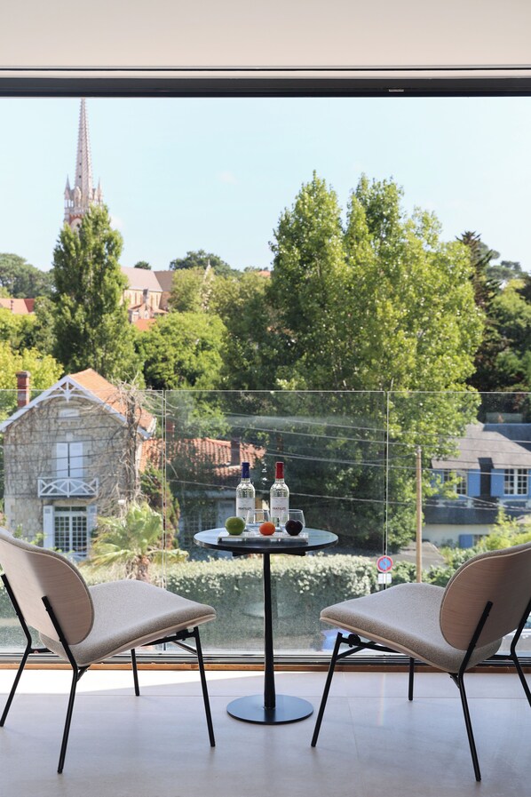 Una habitación moderna con un ventanal grande que da a una ciudad. Hay dos sillones y una mesita redonda con una botella y una copa.