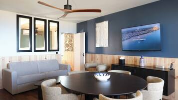 Una sala de estar moderna con una mesa redonda oscura, sillas de mimbre, un sofá gris y un ventilador de techo. Un televisor de pantalla plana montado en la pared muestra una vista panorámica.