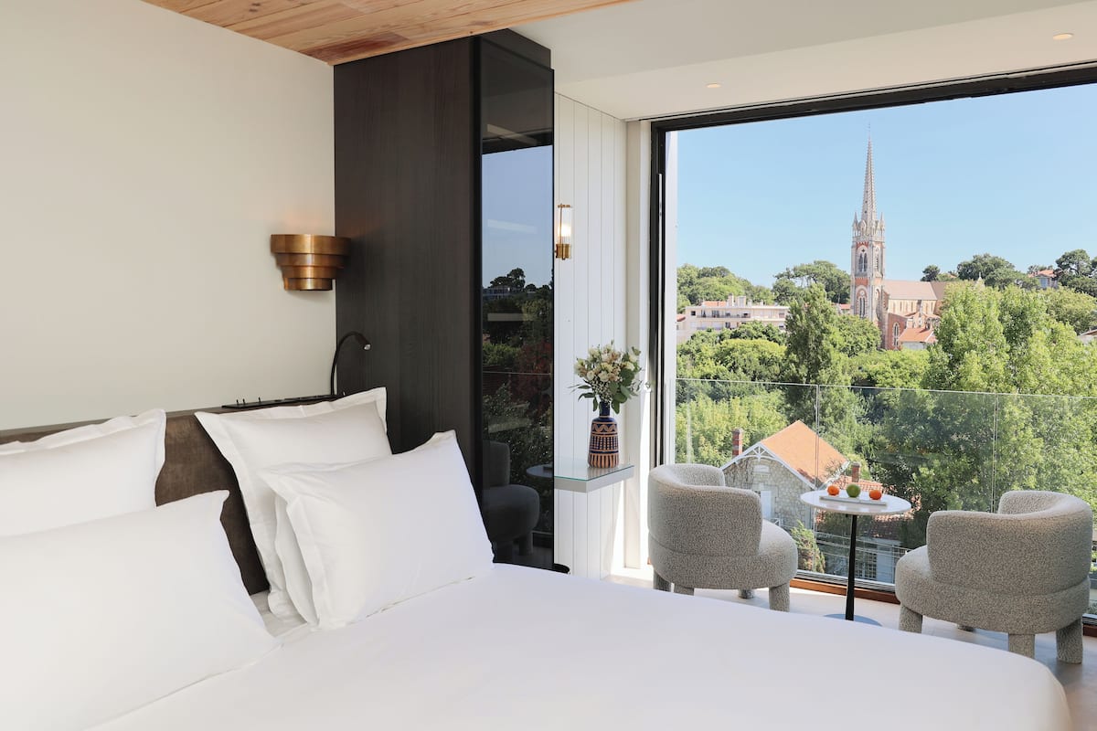 A modern hotel room with a large bed, a seating area with two chairs, and a balcony with a view of a church and greenery.