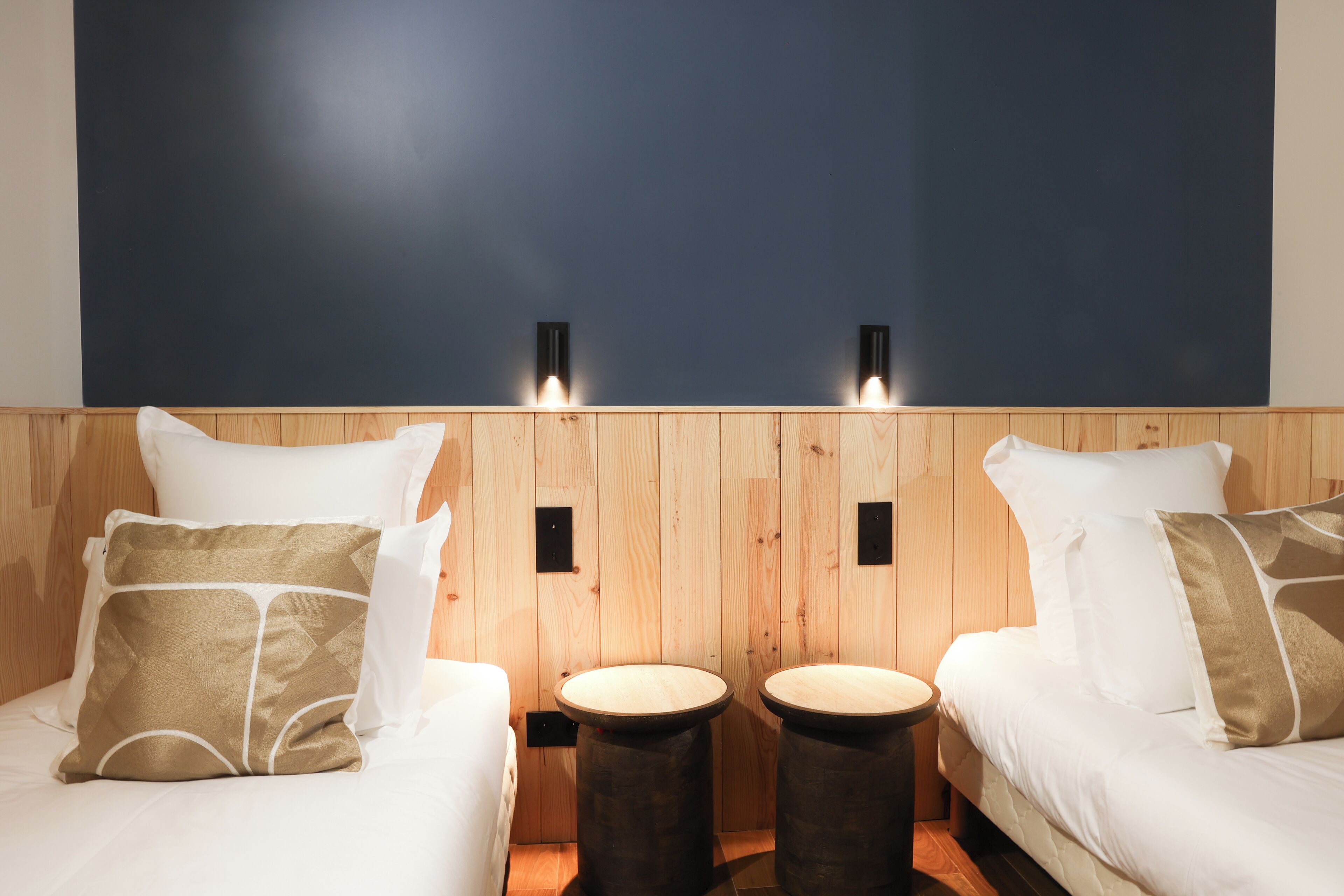 A room with two beds, each with white bedding and beige pillows, and two round wooden stools.