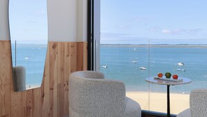 Una habitación moderna con un ventanal grande con vistas al mar, dos sillones tapizados, una mesa redonda con fruta y un espejo de pared original.
