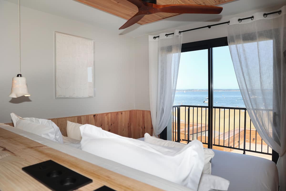 A bedroom with a wooden headboard, a bed with white linens, a bedside table with a lamp, and a view of the beach through a balcony.