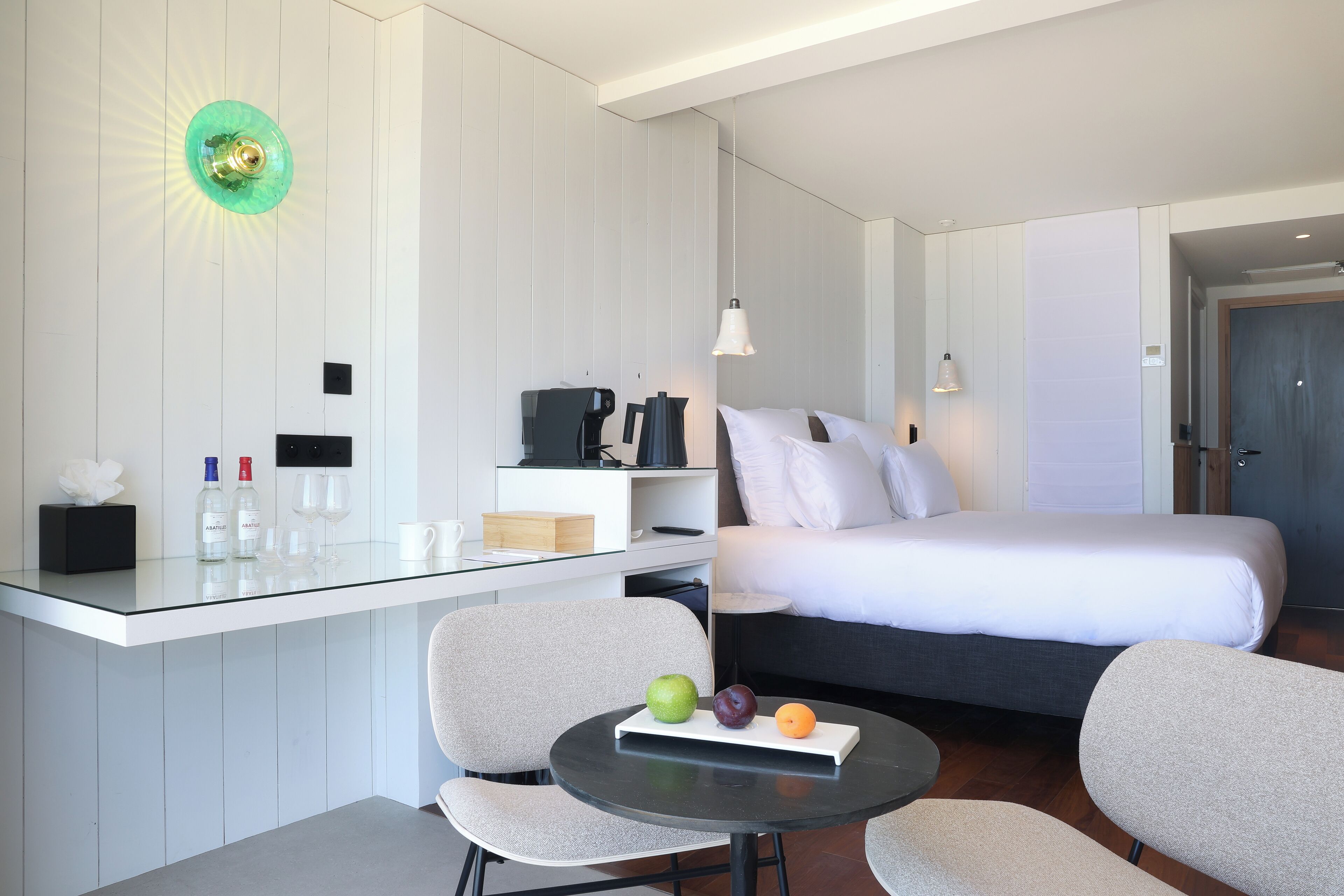 A modern hotel room with a bed, a desk with a glass top, a chair, and a small table with fruit.