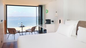 Habitación de hotel con un ventanal grande que ofrece vista al mar, una cama, un escritorio con cafetera y dos sillas.