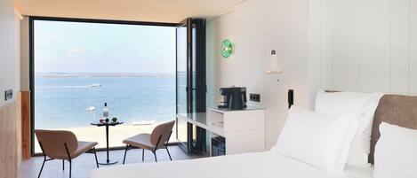 A hotel room with a large window offering a sea view, a bed, a desk with a coffee maker, and two chairs.